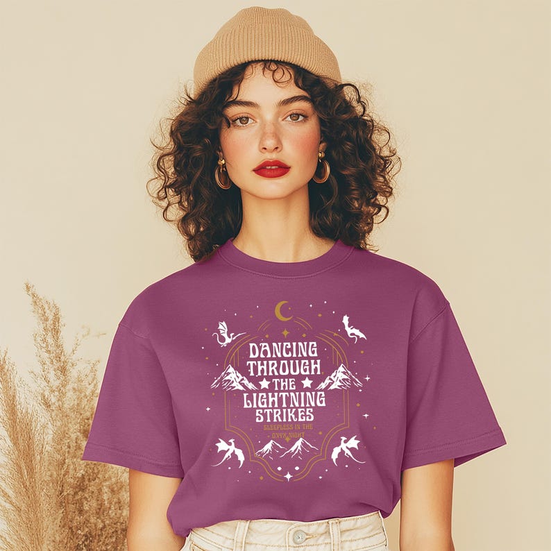Puede incluir: Una camiseta magenta de cuello redondo con el texto "Dancing Through The Lightning Strikes" en un dise&ntilde;o decorativo. El dise&ntilde;o incluye monta&ntilde;as, estrellas y figuras estilizadas. La persona de la imagen lleva un gorro beige.