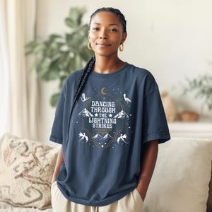 Puede incluir: Una camiseta azul marino con un dise&ntilde;o gr&aacute;fico con el texto "Dancing Through The Lightning Strikes". El dise&ntilde;o incluye una luna creciente, monta&ntilde;as, estrellas y dragones estilizados en blanco y dorado.