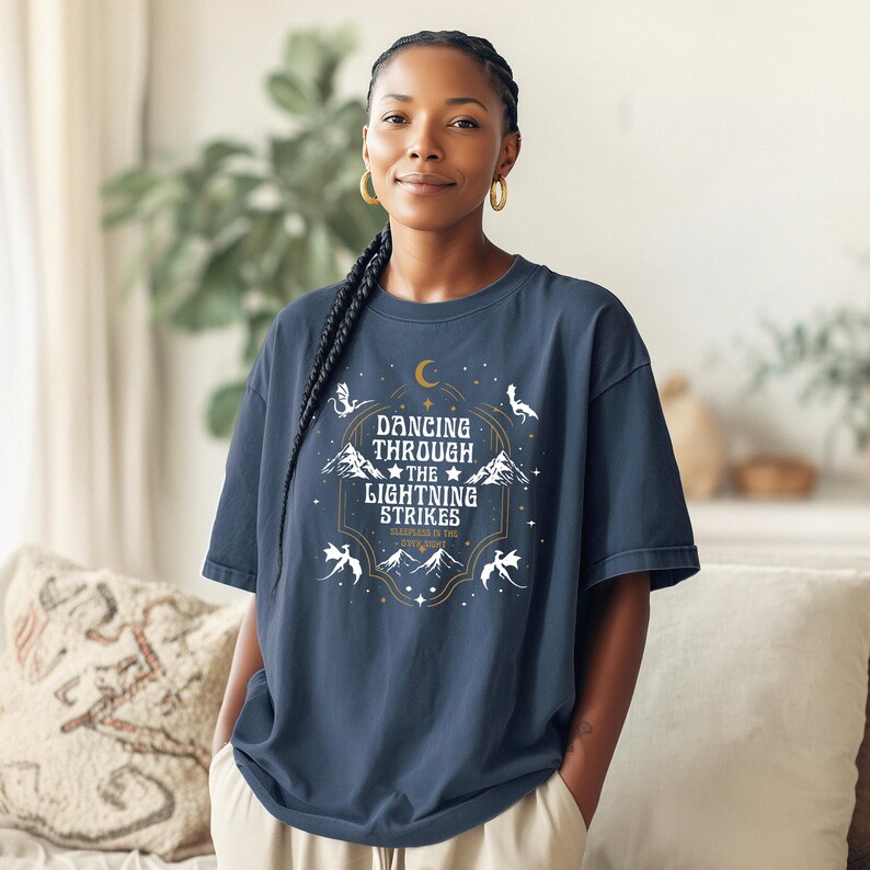 Puede incluir: Una camiseta azul marino con un dise&ntilde;o gr&aacute;fico con el texto "Dancing Through The Lightning Strikes". El dise&ntilde;o incluye una luna creciente, monta&ntilde;as, estrellas y dragones estilizados en blanco y dorado.