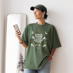 Puede incluir: Camiseta verde oliva con el texto "Dancing Through The Lightning Strikes" en un dise&ntilde;o dorado y blanco. El dise&ntilde;o incluye estrellas, monta&ntilde;as y criaturas m&iacute;ticas. La camiseta es de corte holgado.