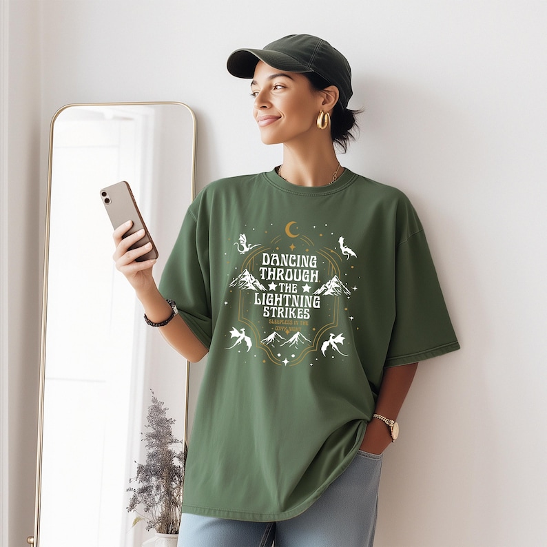 Puede incluir: Camiseta verde oliva con el texto "Dancing Through The Lightning Strikes" en un dise&ntilde;o dorado y blanco. El dise&ntilde;o incluye estrellas, monta&ntilde;as y criaturas m&iacute;ticas. La camiseta es de corte holgado.