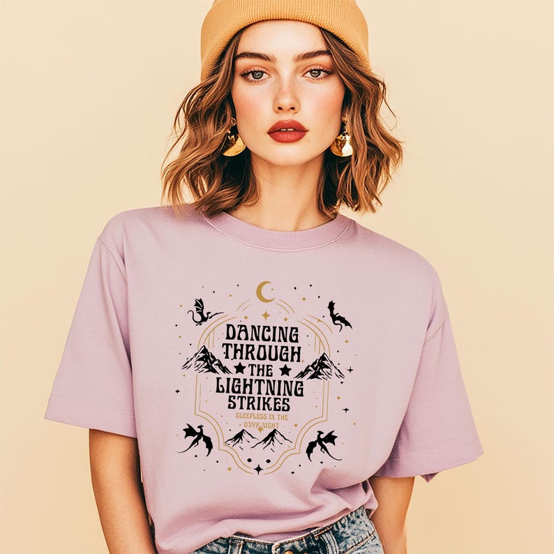 Puede incluir: Camiseta de color lila con un dise&ntilde;o gr&aacute;fico que dice "Dancing Through The Lightning Strikes". El dise&ntilde;o incluye monta&ntilde;as, estrellas y siluetas de dragones. La camiseta tiene mangas cortas y cuello redondo.
