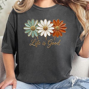 May include: Dark gray t-shirt featuring three daisy flowers in teal, white, and orange with gold accents. The text "Life is Good" is written in gold script below the flowers. The shirt is a casual, comfortable style.