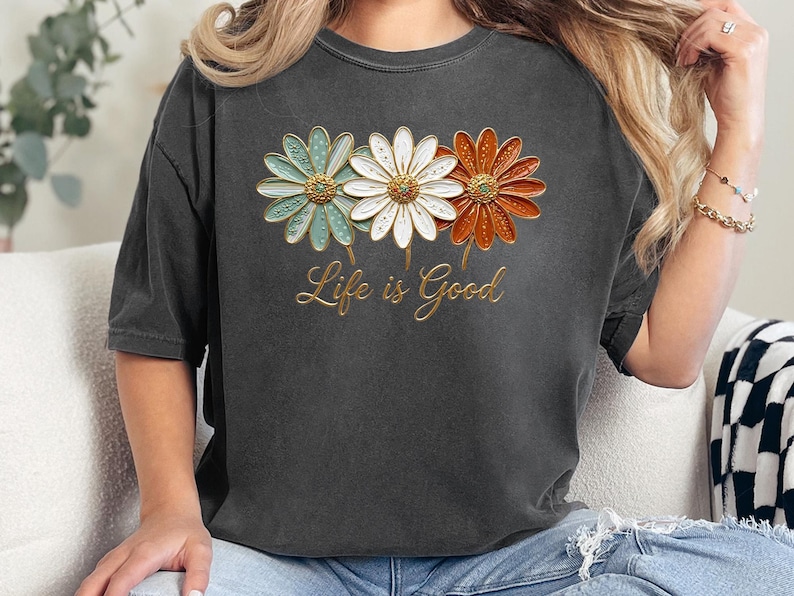 May include: Dark gray t-shirt featuring three daisy flowers in teal, white, and orange with gold accents. The text "Life is Good" is written in gold script below the flowers. The shirt is a casual, comfortable style.