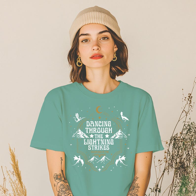 Puede incluir: Una camiseta verde azulado con el texto "Dancing Through The Lightning Strikes" en un dise&ntilde;o blanco y dorado. El dise&ntilde;o incluye monta&ntilde;as, dragones y una luna creciente. La camiseta la lleva una persona.