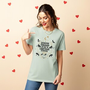 Puede incluir: Camiseta verde claro con el texto "Dancing Through the Lightning Strikes" en un dise&ntilde;o decorativo. El dise&ntilde;o incluye monta&ntilde;as, estrellas y lunas crecientes. La camiseta es de cuello redondo y manga corta.