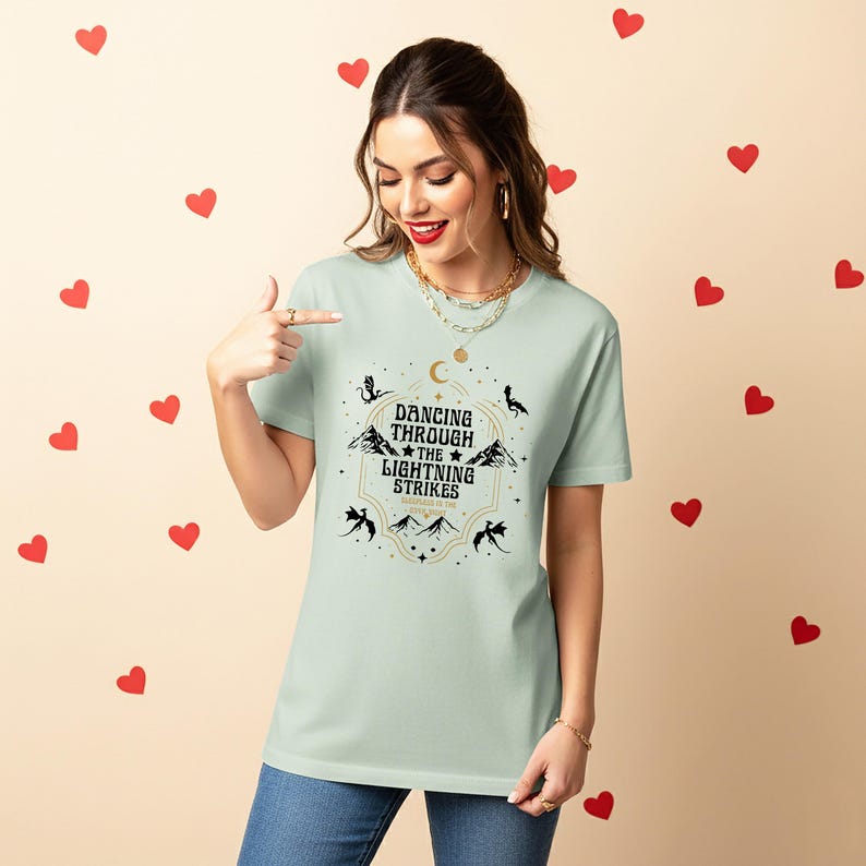 Puede incluir: Camiseta verde claro con el texto "Dancing Through the Lightning Strikes" en un dise&ntilde;o decorativo. El dise&ntilde;o incluye monta&ntilde;as, estrellas y lunas crecientes. La camiseta es de cuello redondo y manga corta.