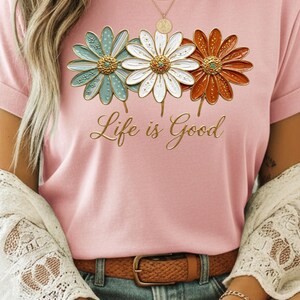 May include: Pink t-shirt featuring three daisy flowers in teal, white, and orange with gold centers. The words "Life is Good" are written in gold script below the flowers. The shirt is worn with a brown belt and blue jeans.