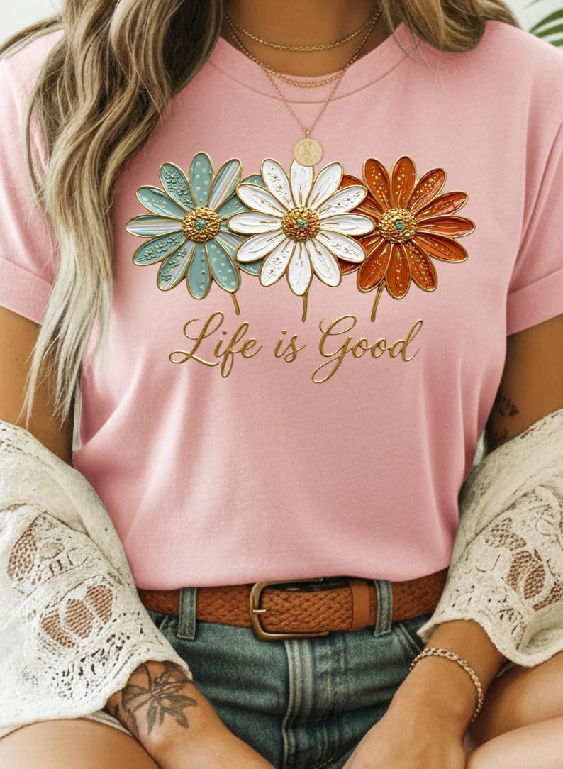 May include: Pink t-shirt featuring three daisy flowers in teal, white, and orange with gold centers. The words "Life is Good" are written in gold script below the flowers. The shirt is worn with a brown belt and blue jeans.