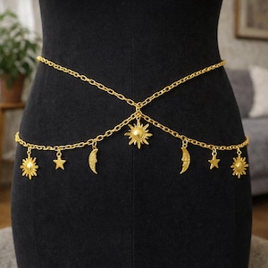 Peut inclure: Chaîne de corps dorée ornée de breloques célestes. La chaîne présente des pendentifs soleil, lune et étoile. La chaîne est drapée autour de la taille, créant un accessoire décoratif.