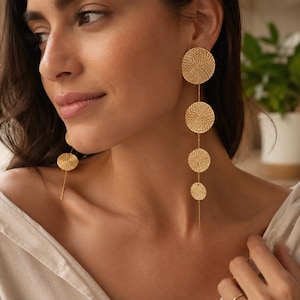 May include: A pair of long gold-toned drop earrings featuring four circular discs of varying sizes arranged vertically. Each disc has a fine, concentric wire-wrapped texture. The earrings hang from the earlobe, creating a linear, dangling silhouette.