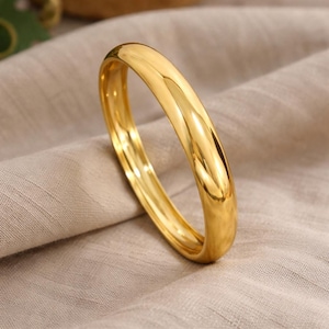 May include: A shiny, gold-colored bangle bracelet. The smooth, rounded design reflects light, creating a bright, polished appearance. The bracelet is displayed on a neutral-toned fabric, highlighting its color and shape. A simple, elegant piece of jewelry.