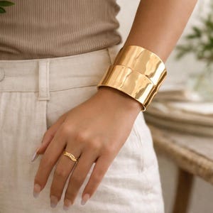 Peut inclure: Un large bracelet manchette doré avec une surface texturée est porté au poignet. Une bague dorée assortie est portée au doigt. Les bijoux se détachent sur un fond neutre, mettant en valeur les pièces audacieuses.