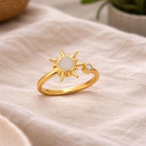 Può includere: Un anello dorato con un design a forma di sole, una pietra bianca centrale e una piccola gemma trasparente sulla fascia. L'anello è posto su un tessuto neutro, evidenziando il suo aspetto luminoso e lucido. Il design del sole è il punto focale.