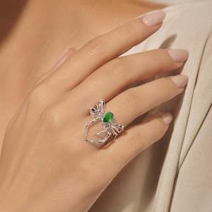 Peut inclure: Une bague argentée en forme d'araignée avec de longues pattes fines qui s'étendent autour du doigt. Le centre de l'araignée présente une unique pierre précieuse verte en forme de poire, sertie solidement dans le corps métallique de l'insecte.