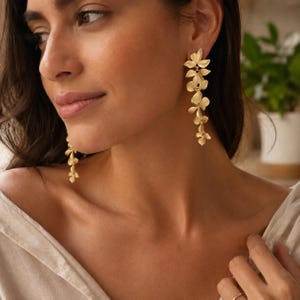 Peut inclure: Une paire de longues boucles d'oreilles pendantes dorées au design floral en cascade. Chaque boucle est composée de multiples éléments en forme de pétales interconnectés qui pendent verticalement, créant un aspect délicat et superposé avec une finition mate.