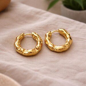 May include: Two gold-coloured hoop earrings with a textured, hammered design. The earrings are set against a neutral-toned fabric background, highlighting their shiny, metallic finish. The earrings are a jewellery item.