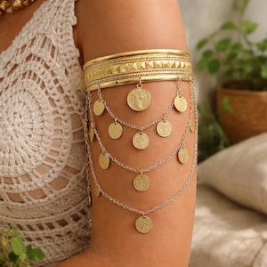 May include: Gold-toned arm cuff with dangling coin charms. The upper band features a geometric design, while multiple chains cascade down, each holding circular pendants. The jewelry is designed to be worn on the upper arm.