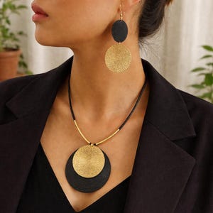 Peut inclure: Un ensemble de bijoux or et noir comprenant un collier et des boucles d'oreilles. Le collier présente un pendentif superposé avec un cercle doré texturé sur un cercle noir. Les boucles d'oreilles reprennent ce design, avec un cercle noir au-dessus d'un cercle doré.