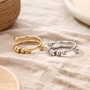 Peut inclure: Deux bagues, une dorée et une argentée, sont présentées sur une surface beige. Chaque bague présente un design à double bande avec de petites perles sphériques. La bague dorée a une finition chaude et brillante, tandis que l'argentée a un aspect métallique poli.