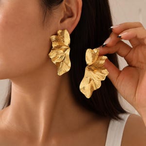 May include: A pair of gold-toned earrings featuring a large, abstract, petal-like design with a textured, rippled surface. The earrings have a metallic finish and a layered, organic shape that extends outward from the earlobe in a fan-like structure.