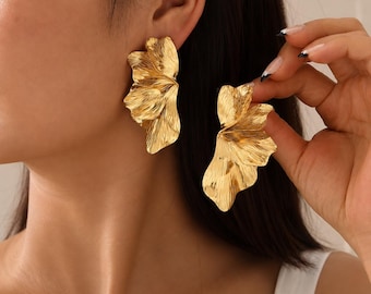 May include: A pair of gold-toned earrings featuring a large, abstract, petal-like design with a textured, rippled surface. The earrings have a metallic finish and a layered, organic shape that extends outwards from the earlobe in a fan-like structure.