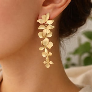 Può includere: Orecchini pendenti floreali color oro. Gli orecchini presentano una cascata di cinque pendenti a forma di fiore, ciascuno con dettagliati disegni di petali. Gli orecchini sono mostrati su uno sfondo color carne.