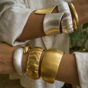 Puede incluir: Una colección de pulseras de metal dorado y plateado. Las pulseras varían en estilo, incluyendo una textura martillada, bandas lisas y un diseño de brazalete ancho. Las joyas se muestran en las muñecas, con una camisa blanca en el fondo.