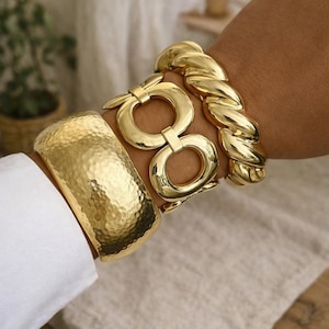 Peut inclure: Plusieurs bracelets dorés. On distingue un jonc martelé, un modèle à maillons et un bracelet torsadé. Les bijoux sont portés au poignet, avec une manche de chemise blanche visible. Ces bracelets sont une pièce forte.