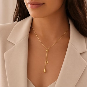 Könnte beinhalten: Eine goldene Lariat-Halskette mit einer dünnen Kette, die durch eine kleine verschiebbare Perle verläuft und in zwei unterschiedlich langen, tropfenförmigen Anhängern endet. Die polierte Metalloberfläche reflektiert das Licht.