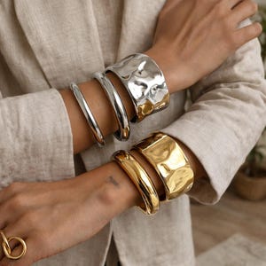 Peut inclure: Bracelets en métal argenté et doré. Les bracelets argentés ont un design lisse et incurvé, tandis que les bracelets dorés ont un aspect texturé et martelé. Les bracelets sont portés aux poignets, mettant en valeur un style de bijoux moderne.