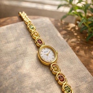 Peut inclure: Montre-bracelet dorée avec un cadran blanc et un bracelet orné de pierres précieuses colorées. La montre présente un cadran ovale et un bracelet délicat et articulé. Les pierres précieuses sont dans les tons de vert, jaune et rouge. La montre repose sur un tissu de couleur neutre.
