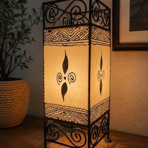 May include: A rectangular table lamp with a black metal frame featuring scrollwork at the top and bottom. The lamp has four off-white panels decorated with black hand-drawn patterns, including geometric lines and central leaf-like motifs with spirals.