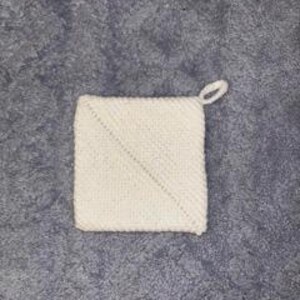 Könnte beinhalten: Ein quadratischer, cremefarbener Topflappen mit einer Schlaufe zum Aufhängen. Der Topflappen hat ein diagonales Design und ist aus einem strukturierten Garn gefertigt. Der Hintergrund ist eine strukturierte, graue Oberfläche.