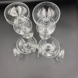 Pode incluir: Seis taças de champanhe transparentes dispostas sobre uma superfície escura. As taças têm uma taça arredondada e uma haste longa. As taças são feitas de vidro transparente e estão vazias.