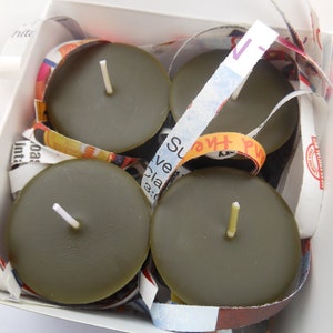 Puede incluir: Cuatro velas de té sin aroma en una caja blanca. Las velas son de color verde oscuro y tienen una mecha blanca. Las velas están rodeadas de trozos de papel de colores.
