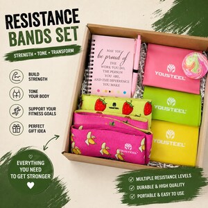 Puede incluir: Un set de regalo de fitness en una caja, que incluye bandas de resistencia, un cuaderno rosa y un bolígrafo. Las bandas son rosas con estampados de plátanos, verdes con fresas y amarillas. El cuaderno tiene el texto "be proud of the work you do".