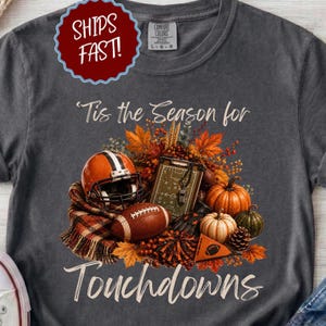 Puede incluir: Una camiseta gris carbón con un gráfico que incluye el texto 'Tis the Season for Touchdowns'. El diseño muestra un casco de fútbol americano, un balón, una pizarra de jugadas, calabazas, hojas de otoño y una bufanda a cuadros en tonos otoñales.
