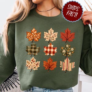 Op de afbeelding: Een olijfgroene sweater met ronde hals en een grafische print van negen esdoornbladeren in een raster. Elk blad heeft een uniek patroon, waaronder ruiten, strepen, bloemmotieven en stippen in de kleuren oranje, rood en crème.