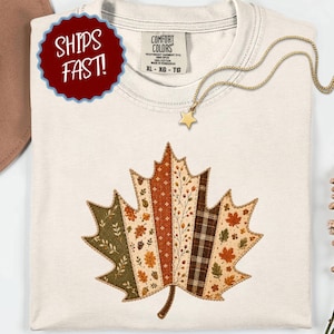 May include: A cream-coloured short-sleeve t-shirt featuring a large, central maple leaf graphic composed of vertical panels with various autumn-themed patterns, including plaid, foliage, and acorns in shades of orange, brown, and olive green.