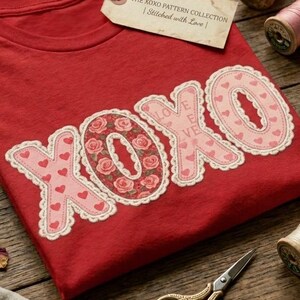 Könnte beinhalten: Ein rotes T-Shirt mit dem Schriftzug XOXO in großen, dekorativen Buchstaben mit Blumen- und Herzmustern, akzentuiert durch eine cremefarbene Spitzenborte. Ein Etikett am Kragen trägt die Aufschrift THE XOXO PATTERN COLLECTION | Stitched with Love |.
