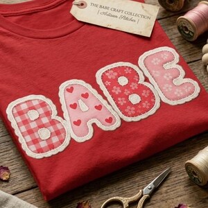 Könnte beinhalten: Ein rotes T-Shirt mit dem Wort BABE in großen, applizierten Stoffbuchstaben mit verschiedenen Mustern wie rotem Vichy-Karo, rosa Herzen und Blumenprints. Ein beiges Etikett trägt die Aufschrift THE BABE CRAFT COLLECTION | Artisan Stitches. Nähzubehör liegt um das T-Shirt herum.