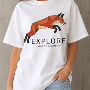 May include: White t-shirt featuring a geometric fox design in shades of orange and brown, with the word "EXPLORE" and the phrase "NATURE'S GEOMETRY" printed below. The fox is depicted mid-jump. The shirt has short sleeves.