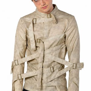 Può includere: Una giacca beige a maniche lunghe con una texture vissuta, caratterizzata da molteplici cinghie in tessuto incrociate fissate con fibbie in metallo color ottone antico sul davanti, sul petto e sulle maniche, con un colletto alto.