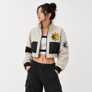 May include: Cropped beige jacket with black accents and a high collar. The jacket features a yellow and black circular emblem on the chest and sleeve. The model is wearing a white and black striped top and black cargo pants.