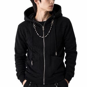 May include: A black zip-up hoodie featuring a silver-toned metal chain draped across the chest, attached to the collar area. The garment includes a hood with black drawstrings, a full-length silver zipper, and two front pockets with silver accents.