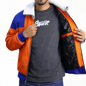 May include: A zip-up jacket featuring a vibrant orange body with blue shoulder and hem accents, a white ribbed collar, and a black quilted interior lining. The jacket is worn over a dark gray t-shirt with the white text "STWD" printed on the chest.