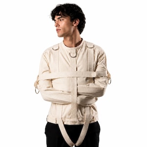 Op de afbeelding: Een beige canvas dwangbuis met lange mouwen, meerdere stoffen banden en zilverkleurige metalen D-ringen. Het kledingstuk is voorzien van een verticale borstband, een horizontale tailleband en extra banden die vanaf de schouders en zijkanten lopen.