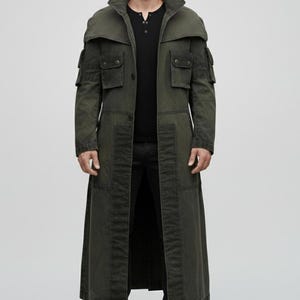 Puede incluir: Un abrigo largo verde oliva con cuello alto y múltiples bolsillos. El abrigo tiene una parte delantera abotonada y un diseño largo y fluido. El modelo lleva una camisa negra y pantalones negros. El abrigo parece estar hecho de una tela duradera.