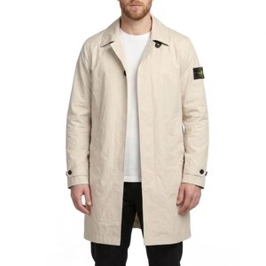 May include: A beige trench coat with a classic collar and button closure. The coat features a small square patch on the upper left sleeve. The model is wearing a white t-shirt and black pants.